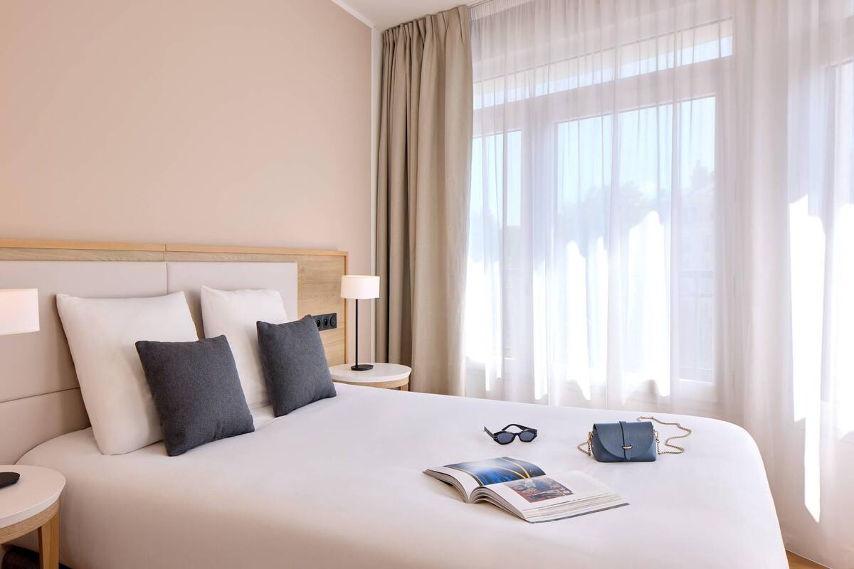 A hotel room with a large bed, two pillows, a nightstand with a lamp, a small table, and a window with sheer curtains.