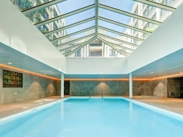 Una piscina interior con techo de cristal, paredes de piedra y una escalera central.