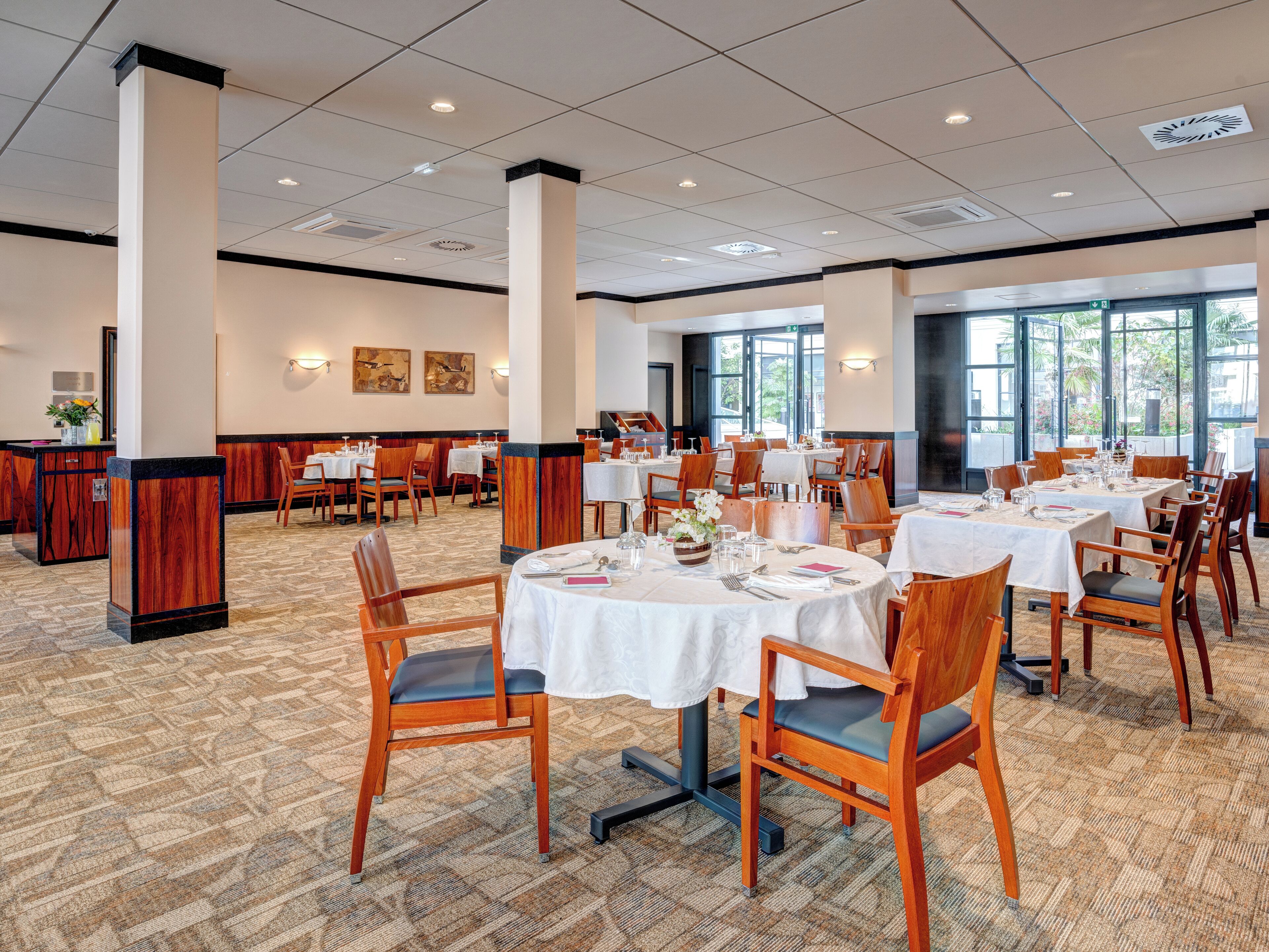 A spacious dining area with multiple round tables, wooden chairs, and a carpeted floor.