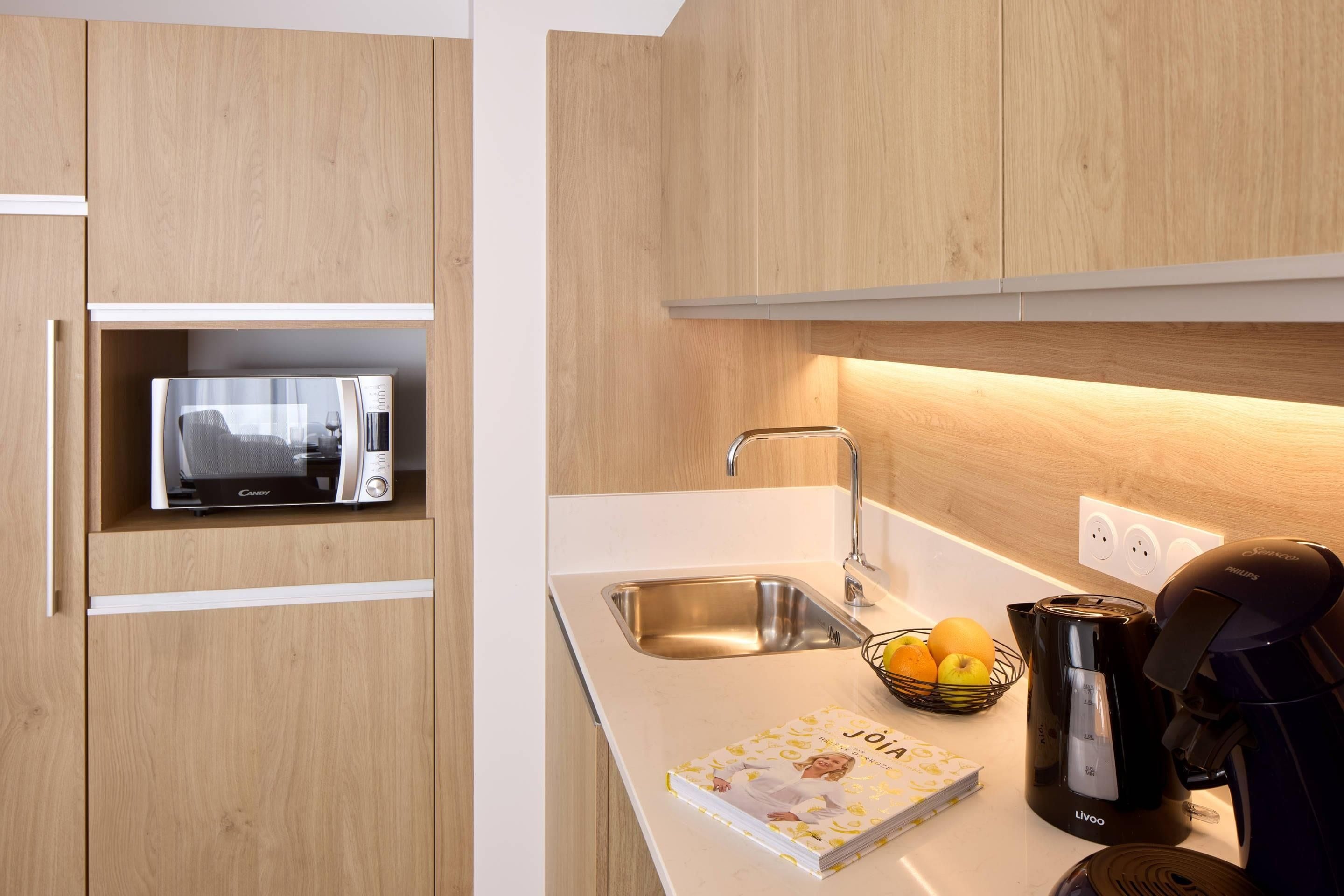 A modern kitchen with wooden cabinets, a built-in microwave, a sink, a coffee maker, and a fruit bowl.