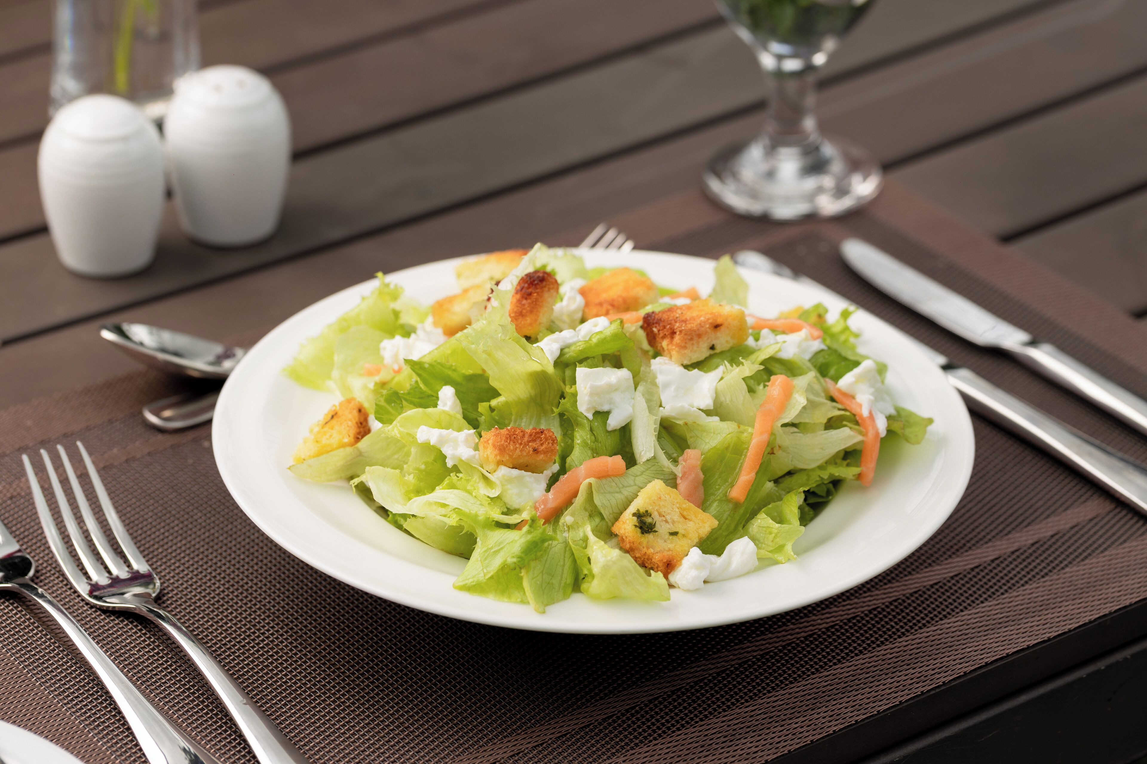 A plate of salad with croutons and shredded carrots on a wooden table with salt and pepper shakers and cutlery.