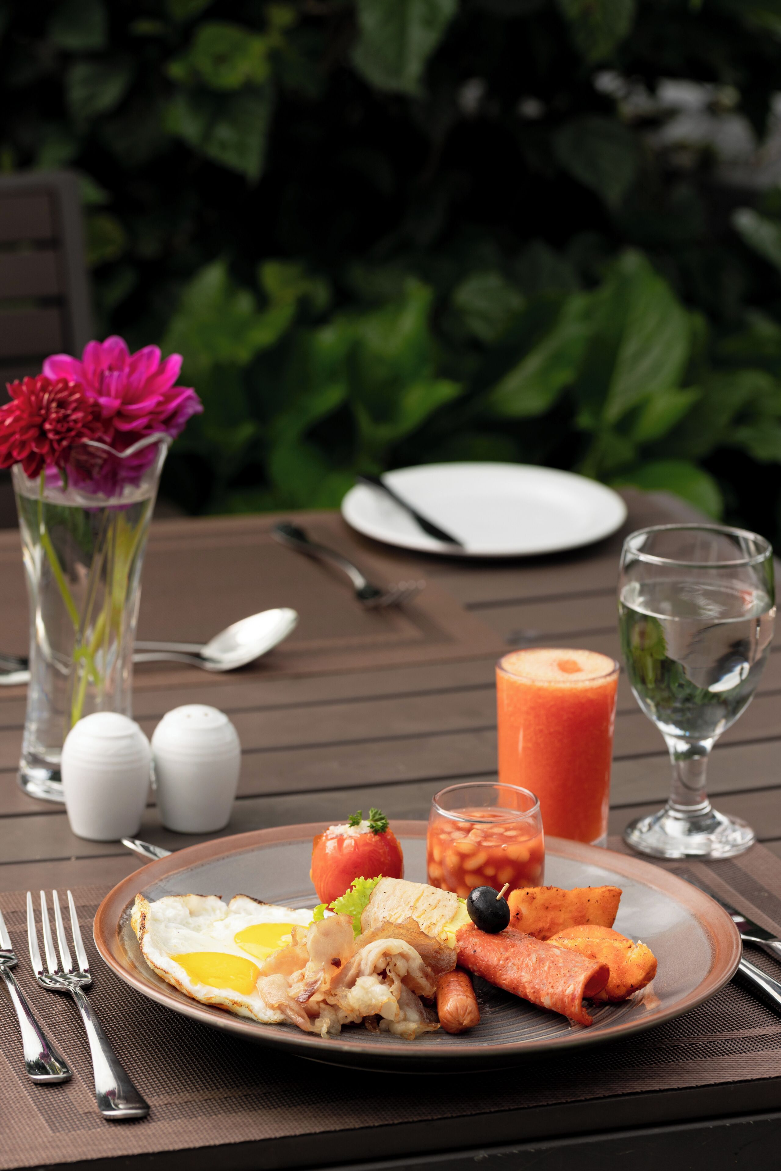 Un plato de desayuno con huevos, salchicha y tomate, acompañado de un vaso de jugo y un florero.