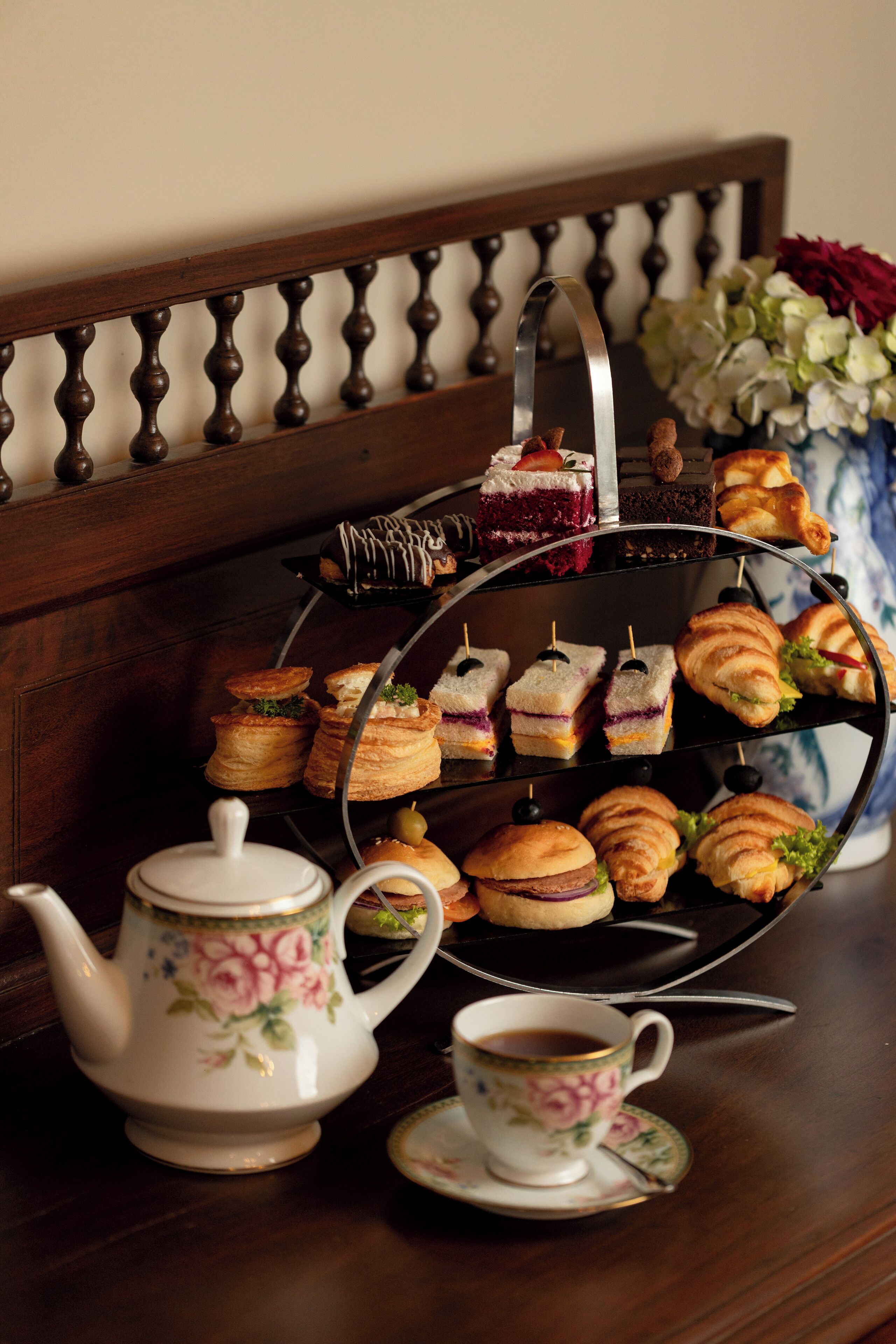 A tea set with a floral teapot and cup, a tiered tray of pastries, and a vase of flowers on a wooden table.