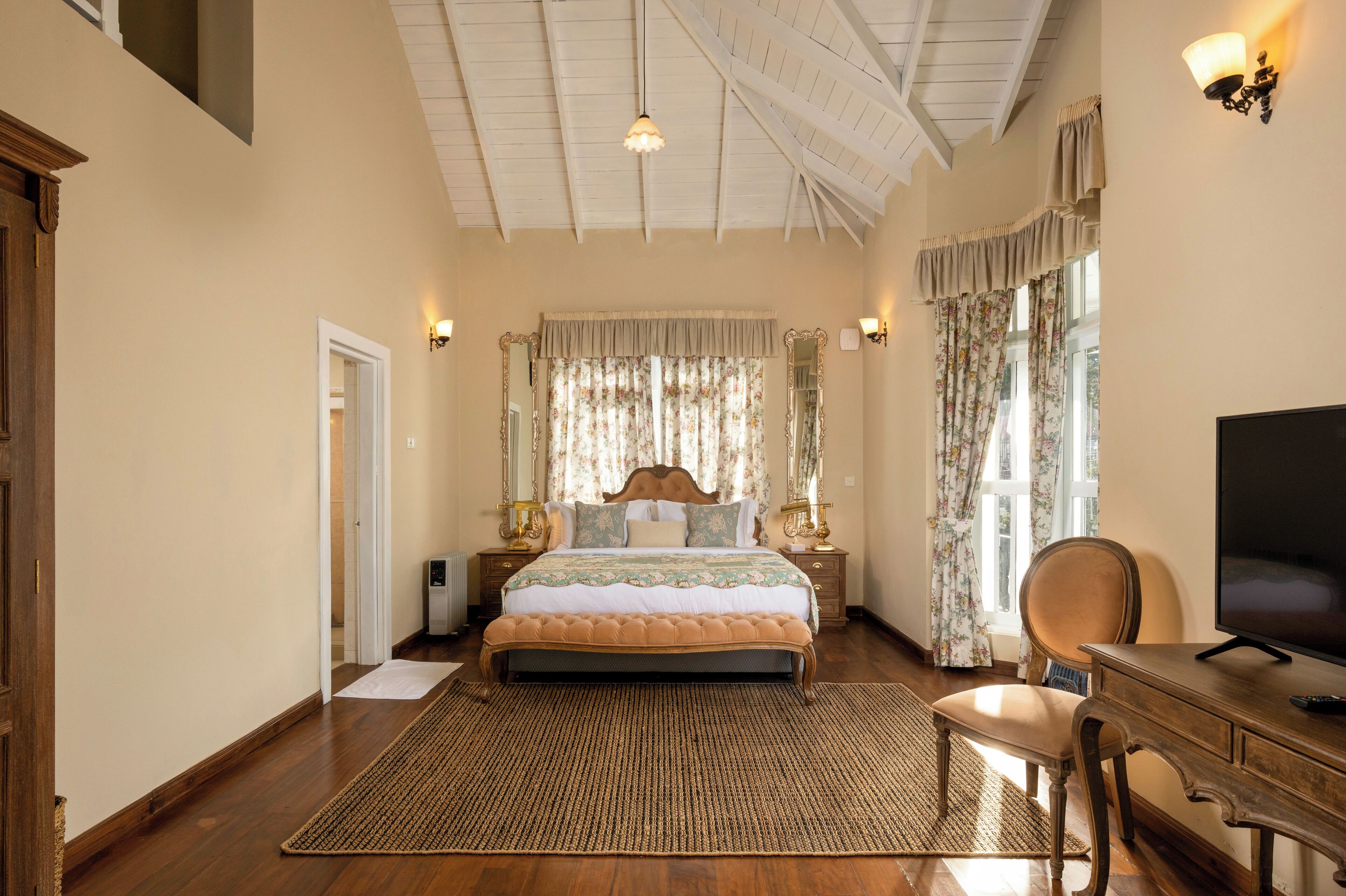 A bedroom with a large bed, wooden headboard, two bedside tables with lamps, a chair, a desk with a TV, and a large window with curtains.