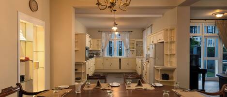 A dining area with a large wooden table set for six, surrounded by wooden chairs. A chandelier hangs from the ceiling, and there are cabinets and a window in the background.
