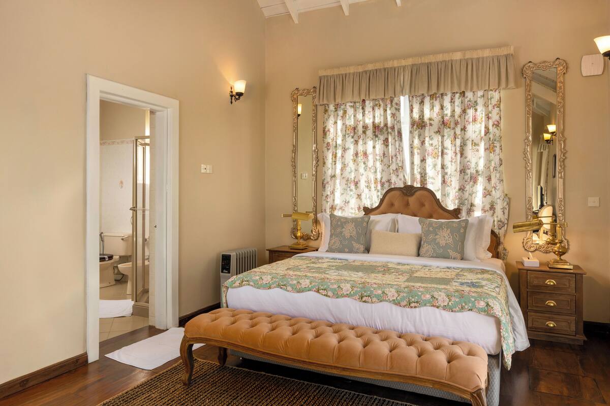 Een slaapkamer met een groot bed, een houten bankje, een nachtkastje met een lamp, een spiegel en een deur naar een badkamer.