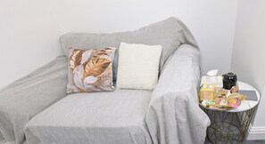 A grey sofa with two pillows, one with a leaf pattern and one plain white, and a small round side table with various items including a black object, a yellow box, and a white container.