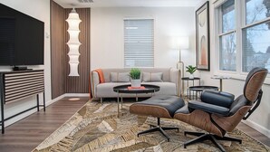 Un salon moderne avec un canapé, une table basse et des fauteuils, un téléviseur et une fenêtre avec des stores.