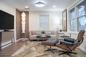 A modern living room with a sofa, coffee table, and chairs, a TV, and a window with blinds.