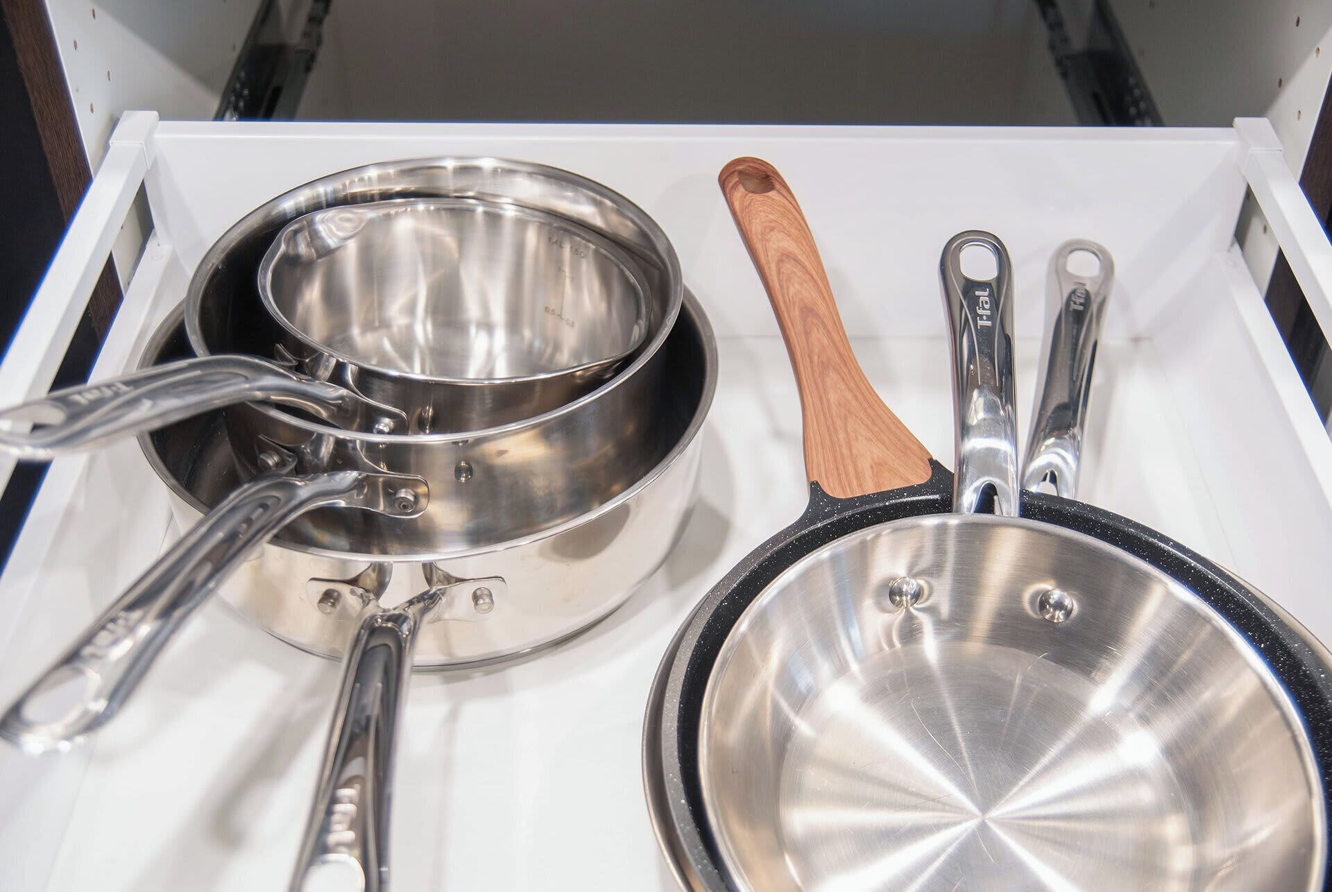 A drawer with kitchen utensils including a pot, a pan, and a wooden-handled utensil.