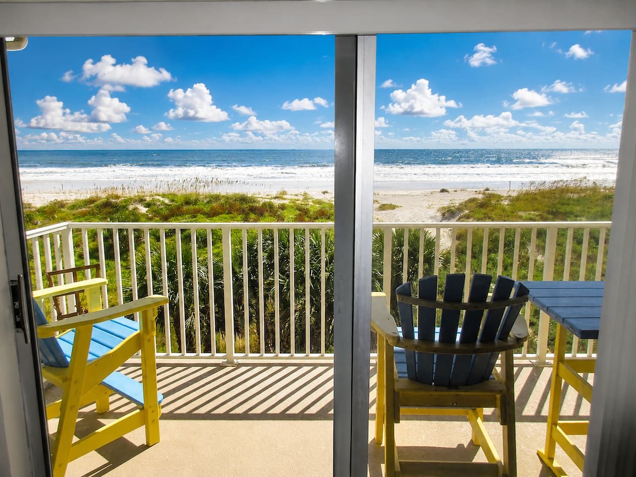 A balcony with a view of the beach, a table, and two chairs.