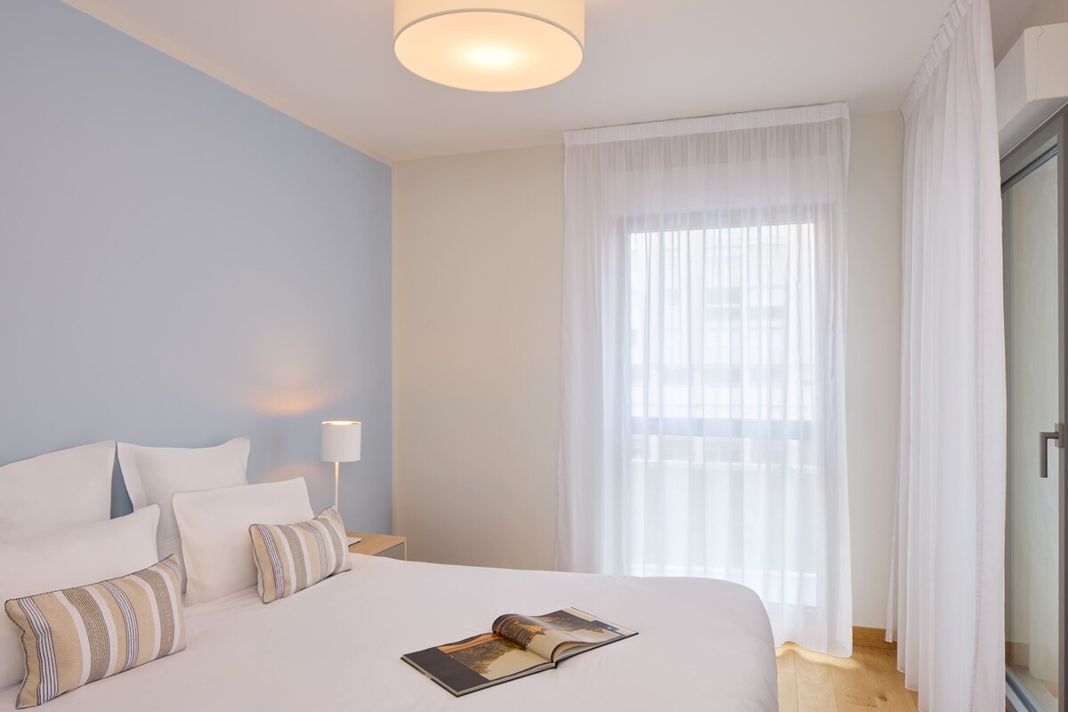 A hotel room with a bed, pillows, a lamp, a window with sheer curtains, and a book on the bed.