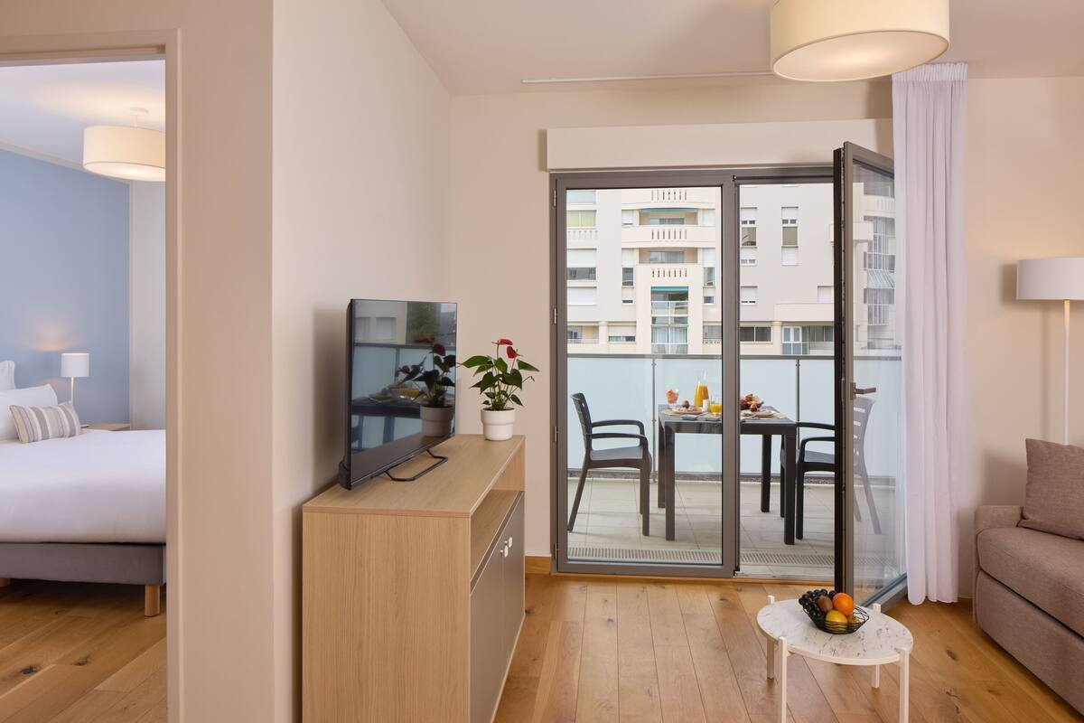 A hotel room with a bed, a TV, a small table with a fruit bowl, a potted plant, and a balcony with a table and chairs.