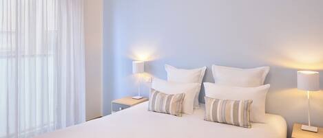 Una cama bien hecha con sábanas blancas, almohadas a rayas y una revista sobre la cama. Hay dos mesitas de noche con lámparas, una ventana con cortinas traslúcidas y vistas del horizonte de la ciudad.