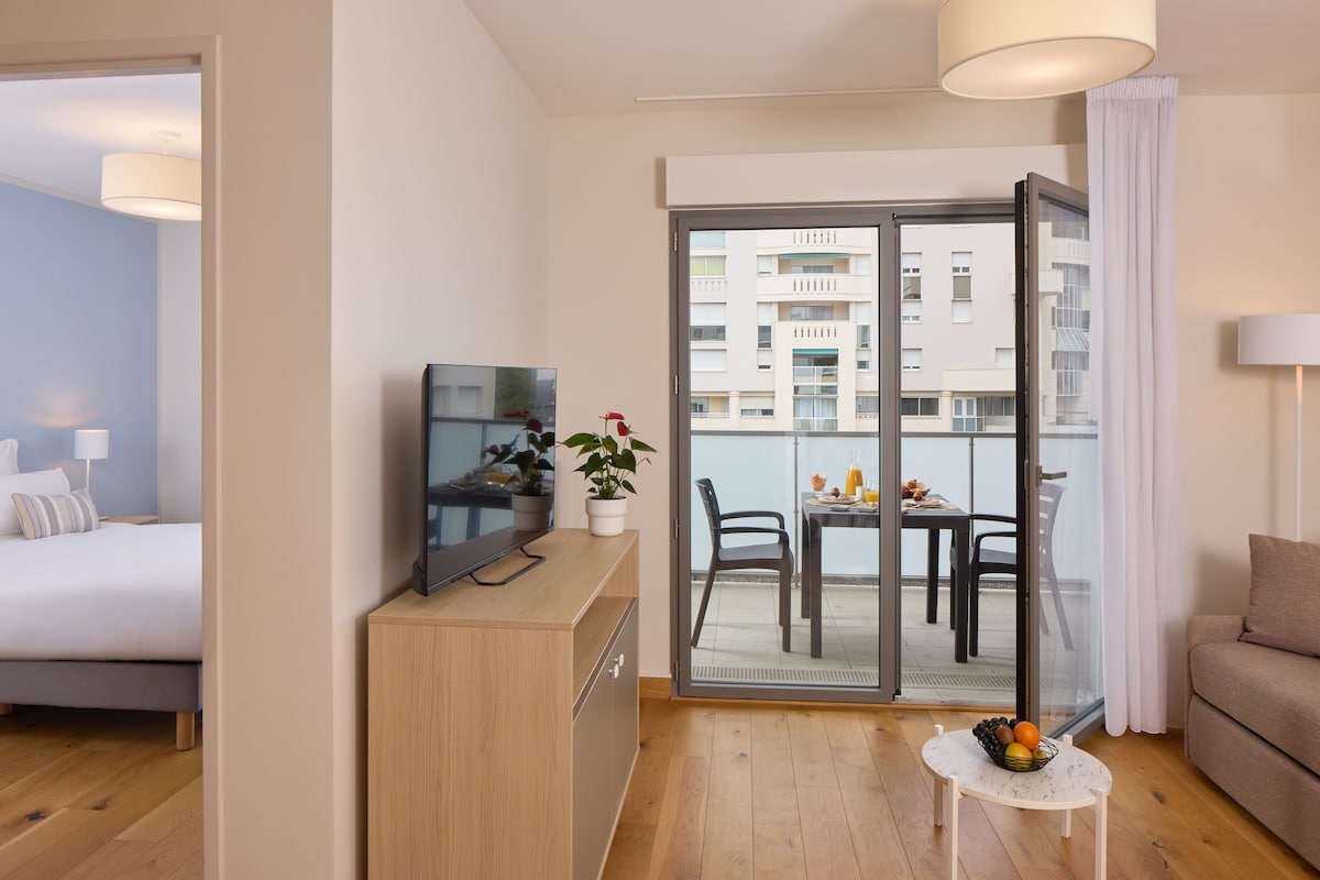 A hotel room with a bed, a TV, a small table with a fruit bowl, a potted plant, and a balcony with a table and chairs.