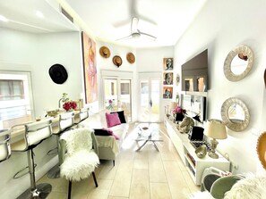 Une pièce lumineuse et bien éclairée, avec un canapé blanc, une table basse en verre et une chaise blanche dotée d’un siège moelleux. Plusieurs chapeaux sont accrochés au mur, et un miroir orné d’un cadre décoratif y est également présent.
