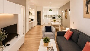Un salon moderne doté d’un téléviseur à écran plat, d’un canapé gris, d’une table basse blanche agrémentée d’une plante, et d’un coin repas à l’arrière-plan.