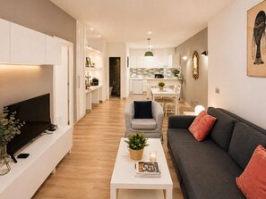 Ein modernes Wohnzimmer mit Flachbildfernseher, einem grauen Sofa, einem weißen Couchtisch mit Pflanze und einem Essbereich im Hintergrund.