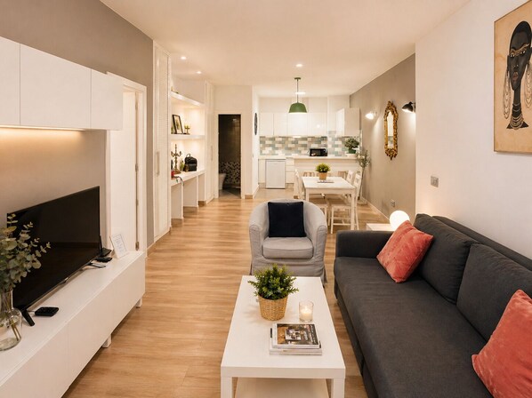 Una sala de estar moderna con un televisor de pantalla plana, un sofá gris, una mesa de centro blanca con una planta y un comedor al fondo.