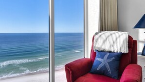 A red armchair with a white throw blanket and a blue pillow with a starfish design, positioned by a large window overlooking a beach and ocean. - Tower 2, three bedroom! Great Views!! FREE BEACH CHAIRS! (Panama City Beach)
