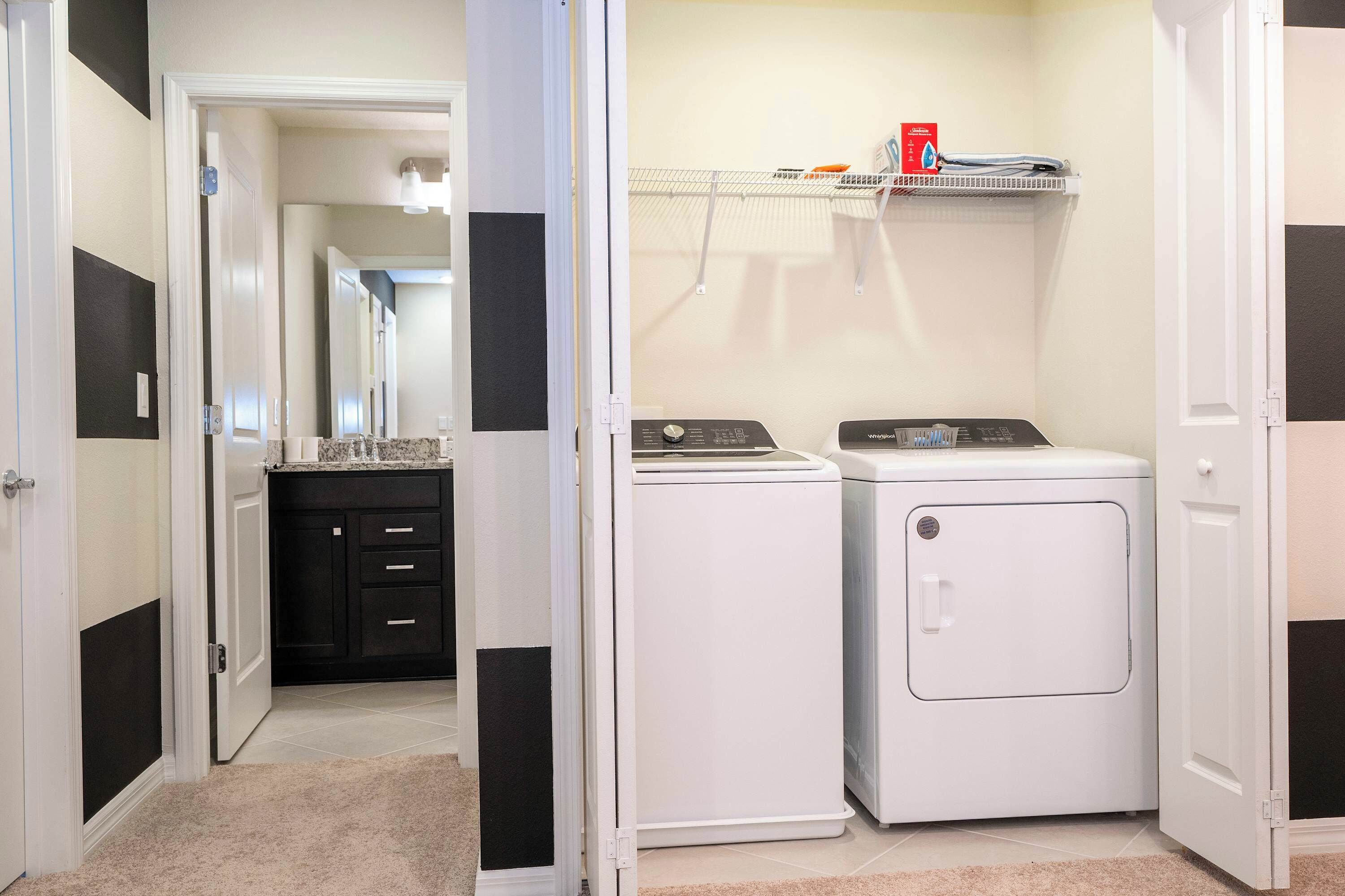 Une buanderie avec deux machines à laver et deux sèche-linge blancs, un mur à rayures noires et blanches, et une étagère contenant des produits de lessive.
