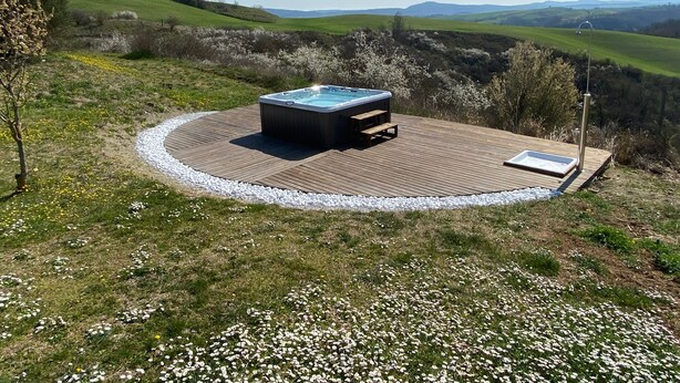 A hot tub on a wooden deck surrounded by a white gravel border, set against a backdrop of green hills and a clear sky. - Corbezzolo (Asciano)