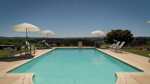 A rectangular swimming pool with a curved edge, surrounded by beige tiles. There are four white umbrellas providing shade, and several lounge chairs are arranged around the pool. - Corbezzolo (Asciano)