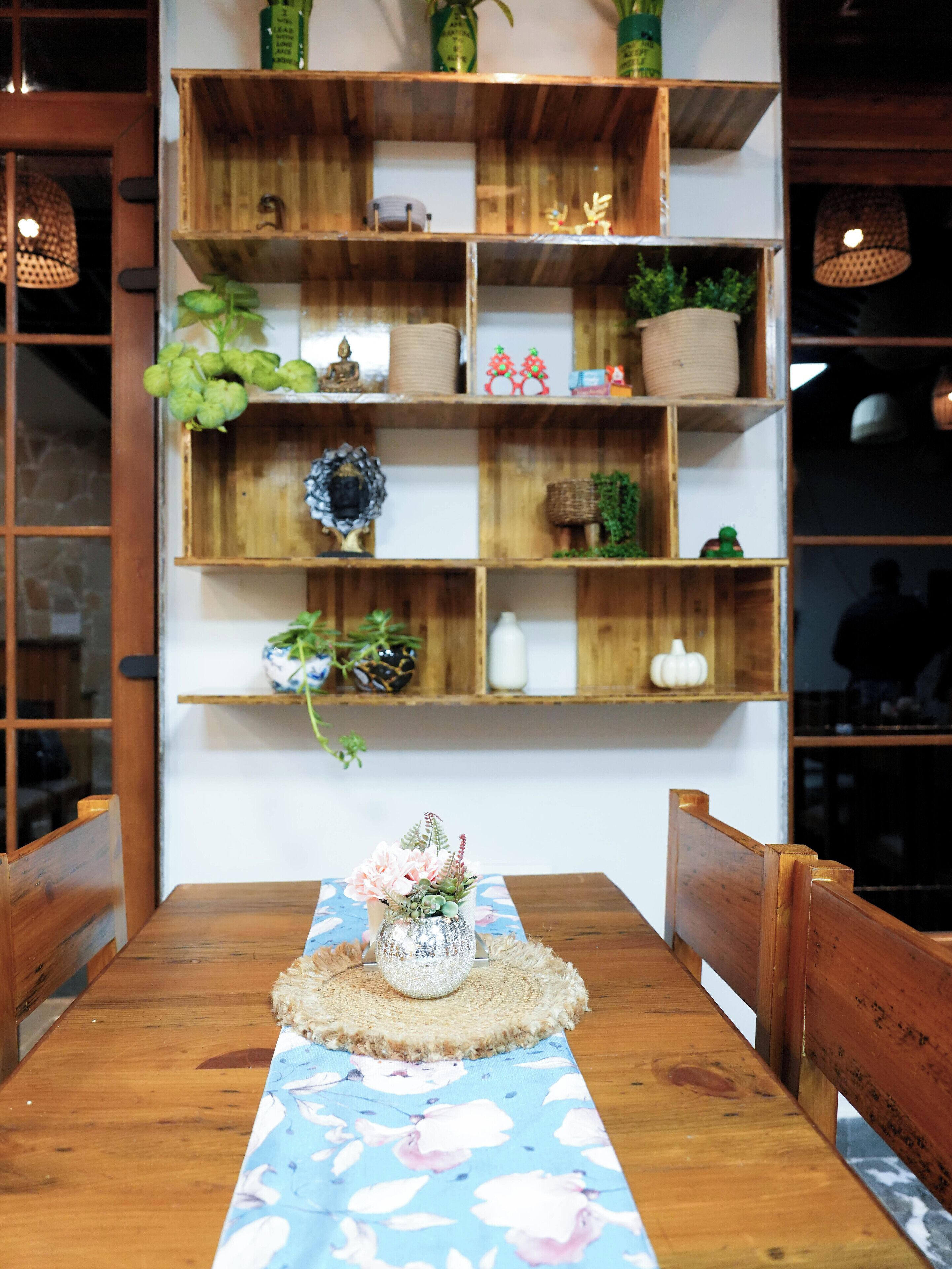 A wooden dining table with a floral table runner, surrounded by wooden chairs. Above the table, there are wooden shelves with various decorative items and plants.