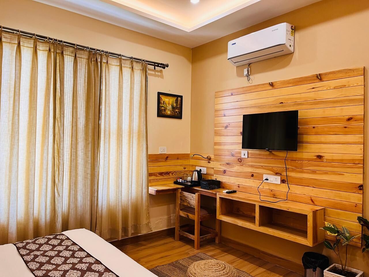 A hotel room with a wooden wall-mounted TV, a small desk with a kettle, and a window with curtains.