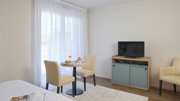 Habitación de hotel con cama, una mesita redonda con sillas, un televisor en un mueble y una ventana con cortinas de gasa.