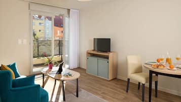 Ett modernt vardagsrum med en soffa, ett matbord dukat för frukost, en tv på ett tv-bord och en balkong med utsikt över byggnader.