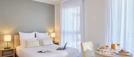 Una habitación de hotel con una cama bien hecha, una mesa de comedor con alimentos para el desayuno y un ordenador portátil sobre la cama.