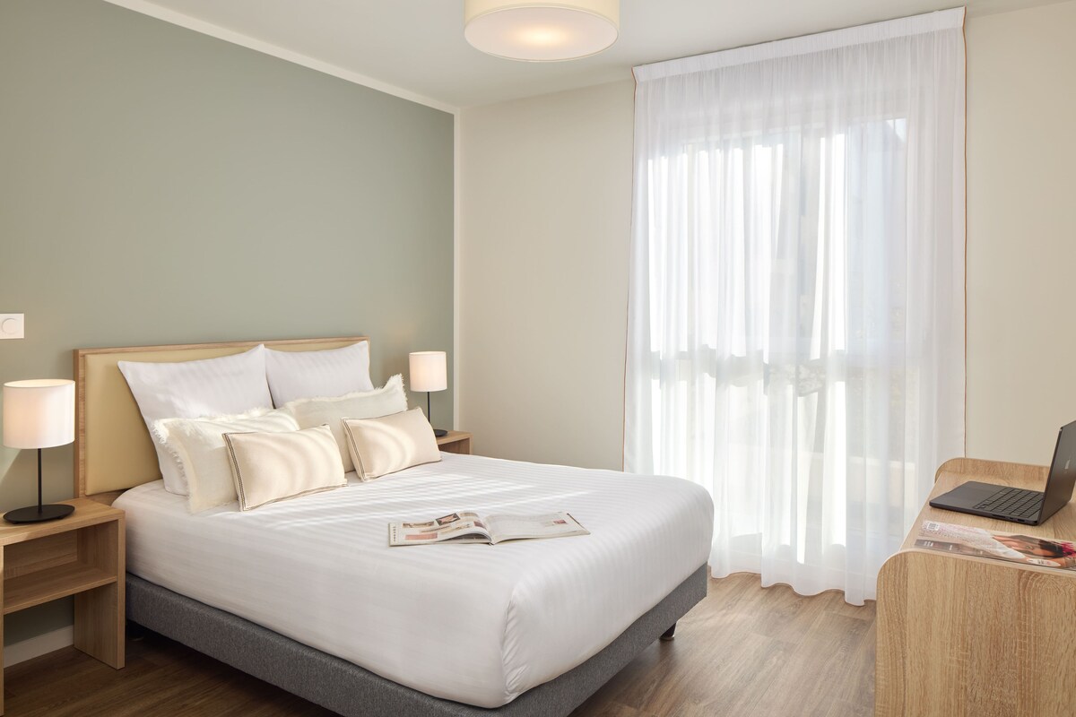 Une chambre d’hôtel avec un lit, des lampes de chevet, un bureau avec un ordinateur portable et une fenêtre avec des rideaux légers.