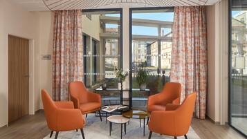 Una habitación moderna con dos sillones naranja, una pequeña mesa y ventanas amplias con cortinas estampadas.