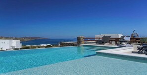 A clear blue pool with lounge chairs and a stone wall, overlooking a coastal landscape. - Luxury White Villa w Private Pool in Mikonos (Mykonos)