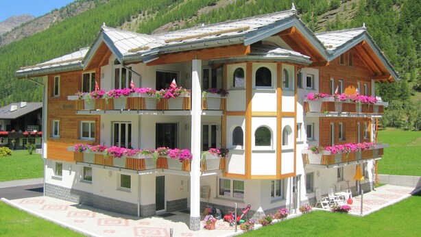 A multi-story hotel with balconies adorned with flowers, set against a mountainous backdrop. - Alpi Retreat by Saas Grund (Saas Grund)