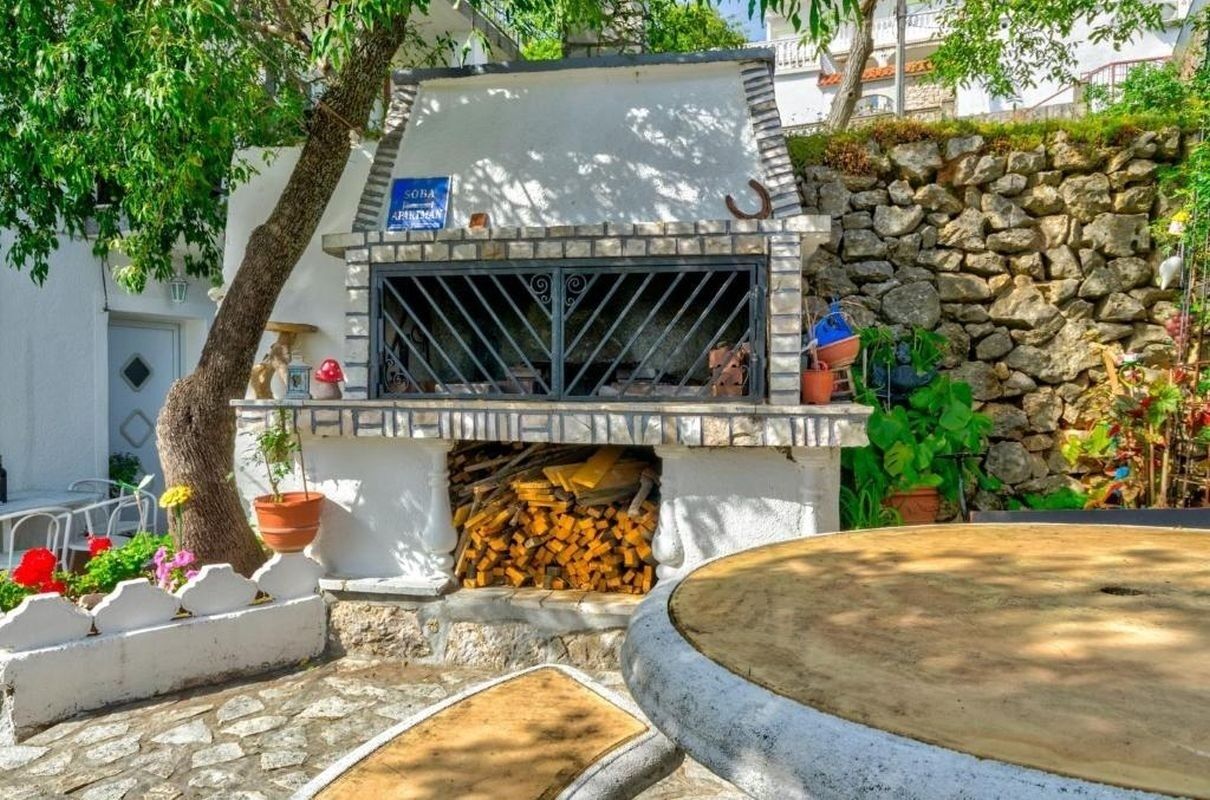 A stone wall with a rustic outdoor grill, surrounded by potted plants and a tree.
