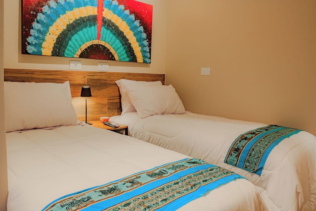 A hotel room with two beds, a wooden headboard, a nightstand with a lamp, and a colorful abstract painting on the wall.