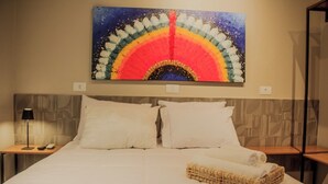 A hotel room with a large bed, two white pillows, a nightstand with a lamp, and a wall-mounted painting of a colorful arch. - Che Hermano Suítes (Foz do Iguaçu)
