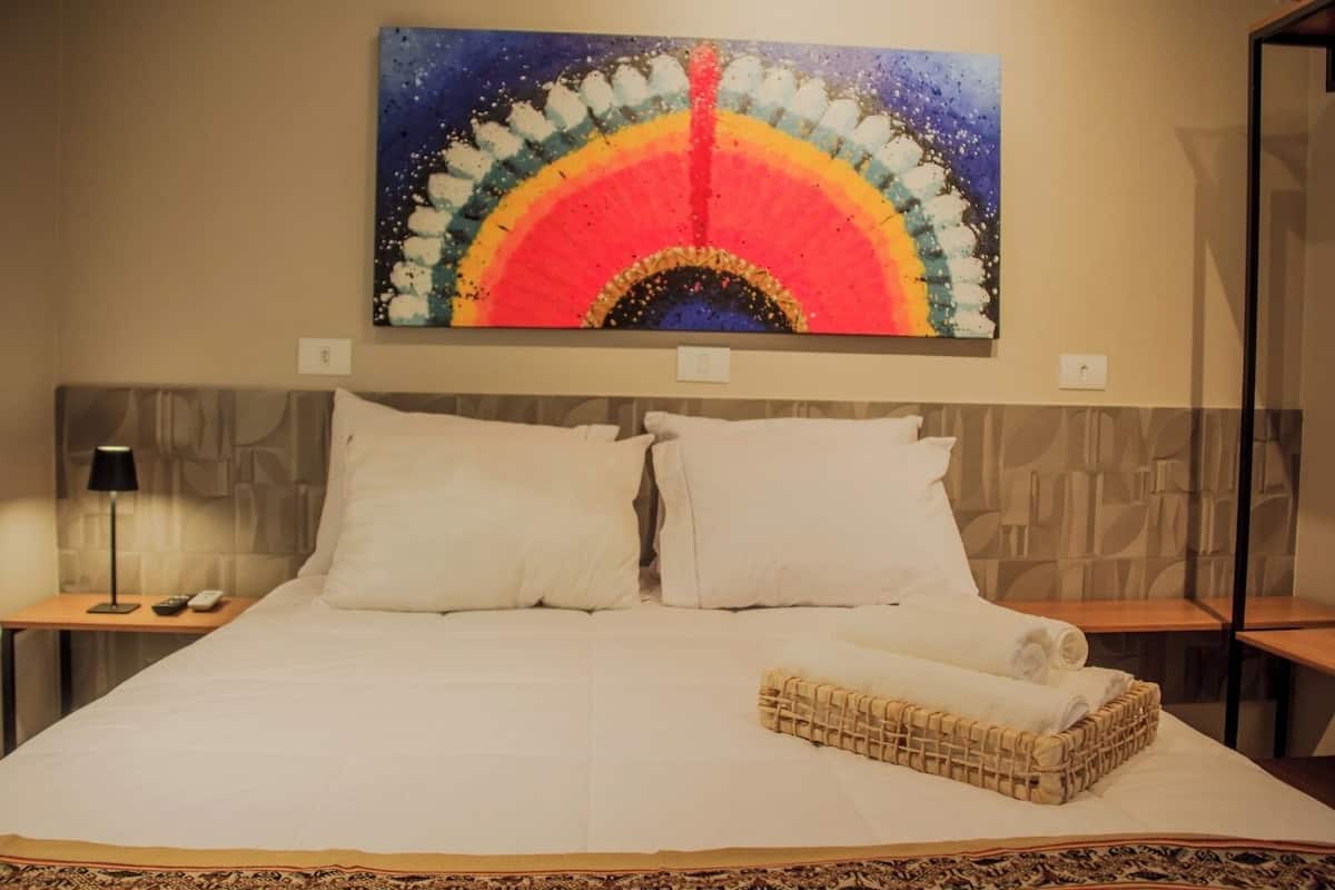 A hotel room with a large bed, two white pillows, a nightstand with a lamp, and a wall-mounted painting of a colorful arch.