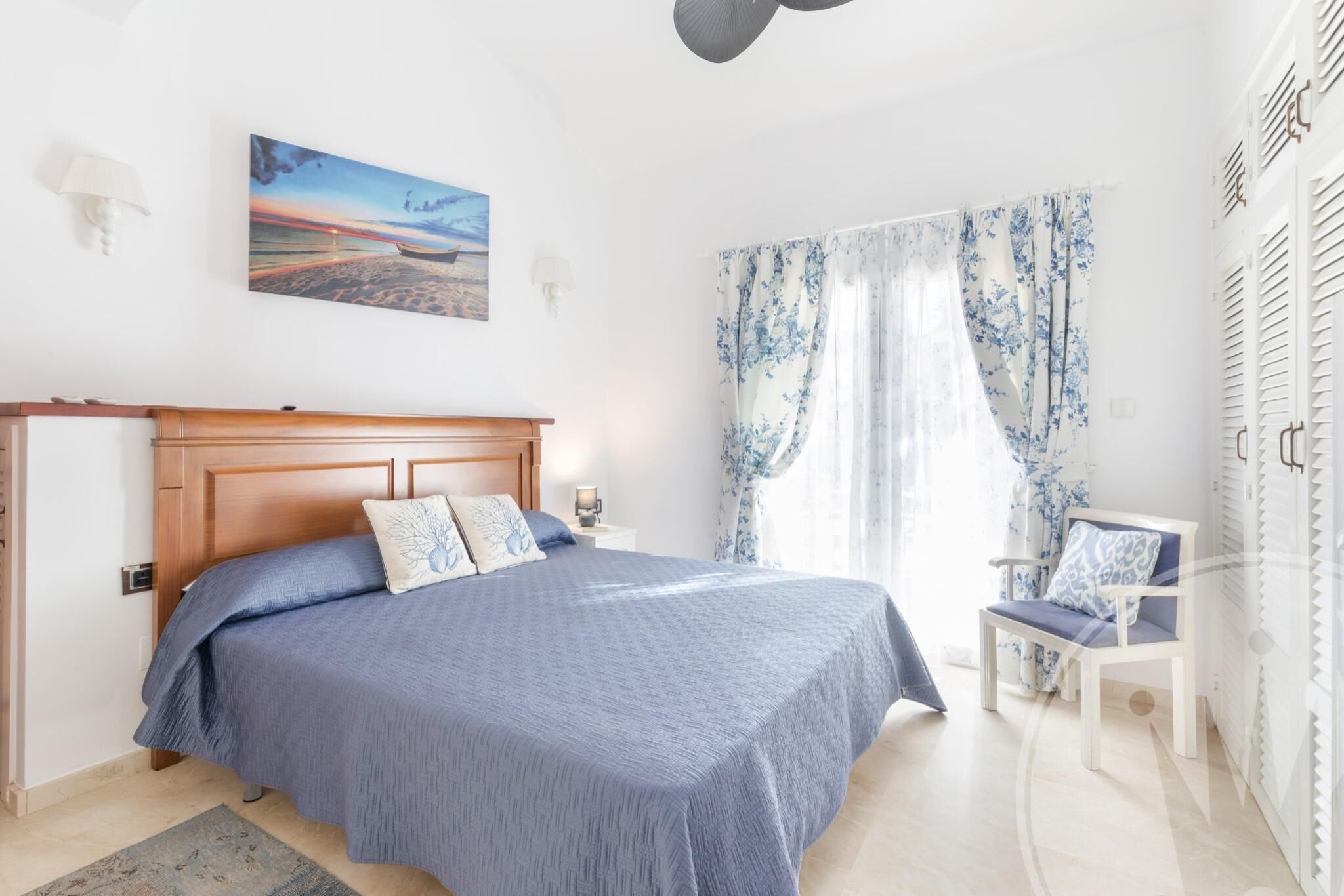 A bedroom with a wooden bed, a nightstand, a chair, and a window with floral curtains.