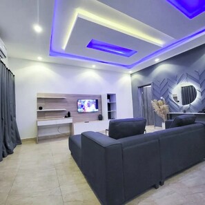 A modern living room with a flat-screen TV, built-in shelves, a dark sofa, and a decorative mirror. - Urban Edge Comfort Apartment (Lokoja)