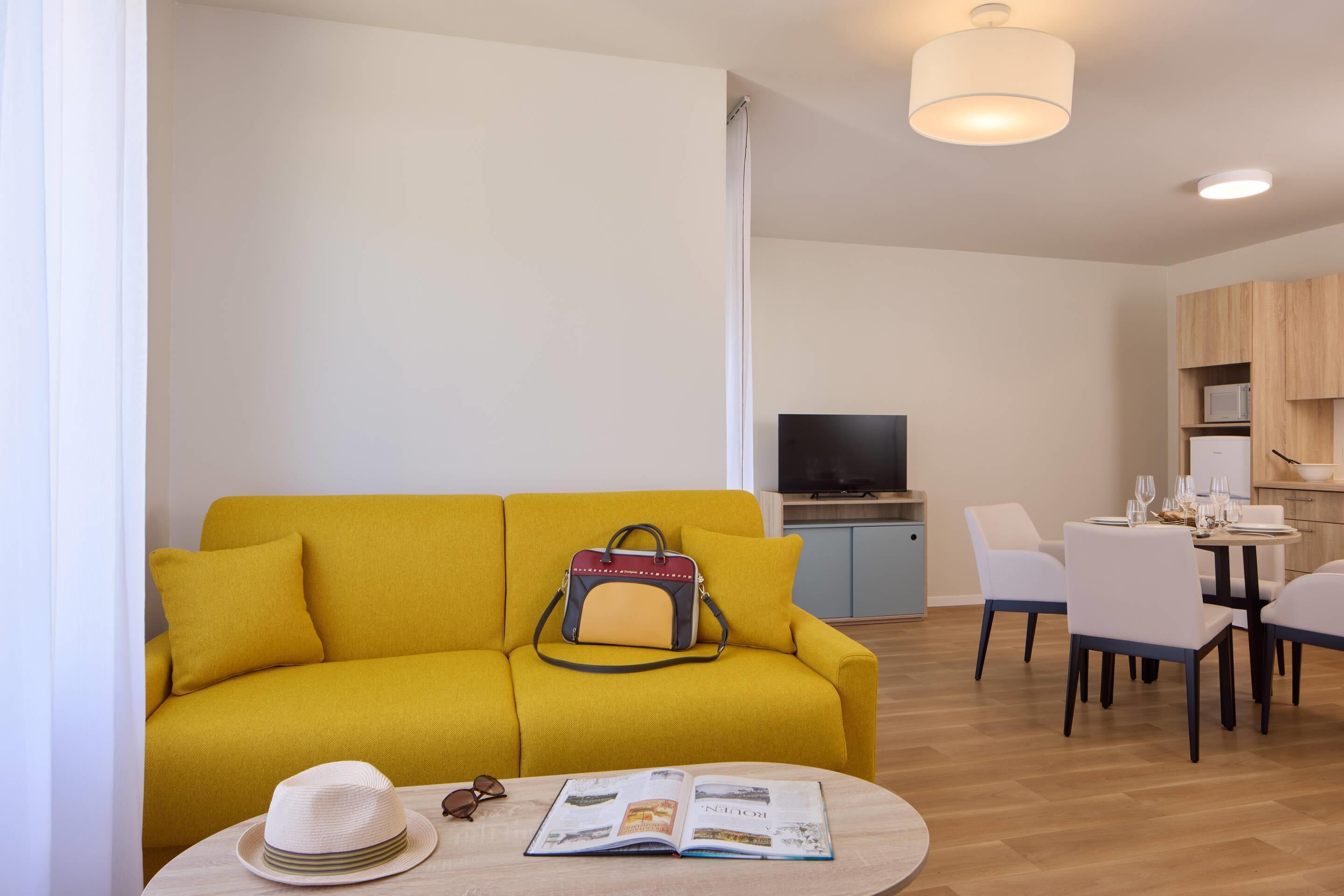Una sala de estar moderna con un sofá amarillo, una zona de comedor con una mesa para cuatro, y una cocina al fondo.