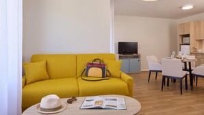 Ein modernes Wohnzimmer mit einem gelben Sofa, einem Essbereich mit Tisch für vier Personen und einer Küche im Hintergrund.
