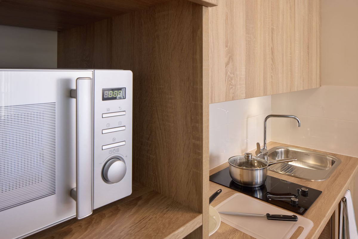 A compact kitchen with a microwave, a digital clock, a sink, and a cutting board.
