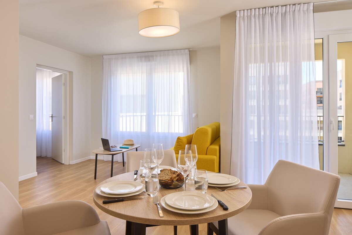 Un coin repas avec une table ronde dressée pour quatre personnes, un ordinateur portable posé sur une petite table d’appoint et un fauteuil jaune en arrière-plan.