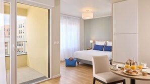 Ein Hotelzimmer mit Bett, einem Esstisch mit Frühstücksangeboten, einem Stuhl und Blick auf die Stadt durch eine Balkontür.