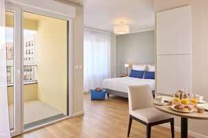 Habitación de hotel con cama, una mesa de comedor con alimentos para el desayuno, una silla y vista a la ciudad a través de una puerta al balcón.