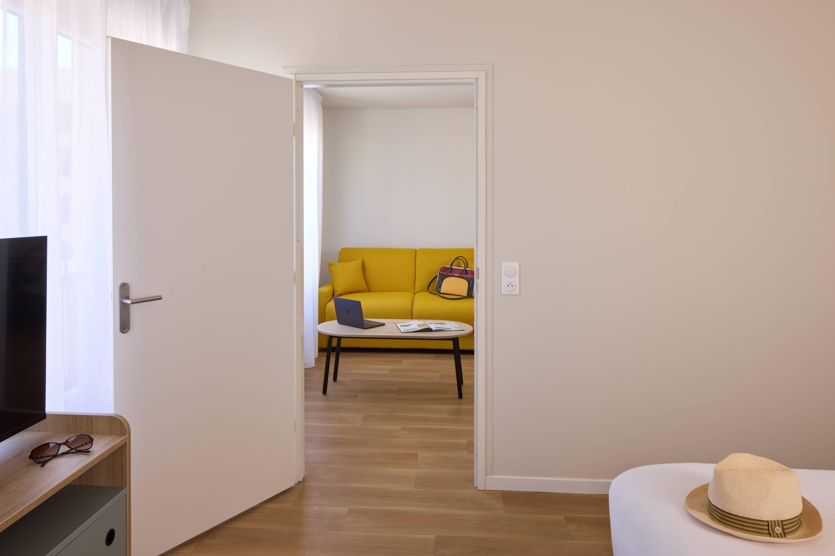 Una habitaci ón con piso de madera, un sofá amarillo, una mesita redonda con una computadora portátil y un libro, y una puerta blanca.