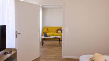 A room with a wooden floor, a yellow sofa, a small round table with a laptop and a book, and a white door.